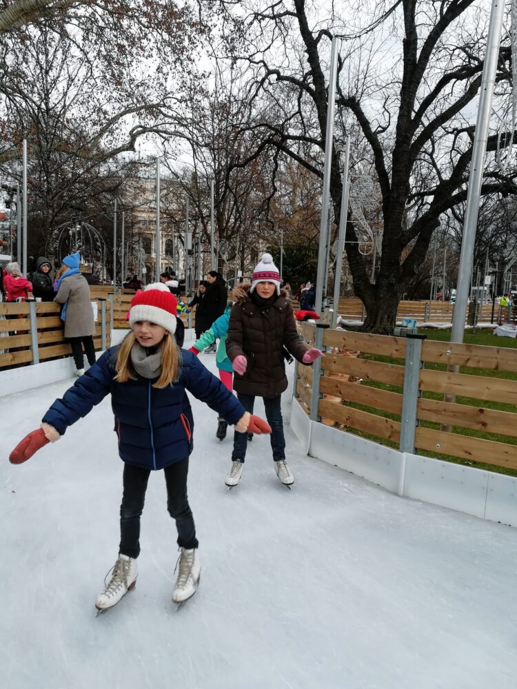 Two girls ice skating in Vienna's Rathaus Park on an overcast winter day.