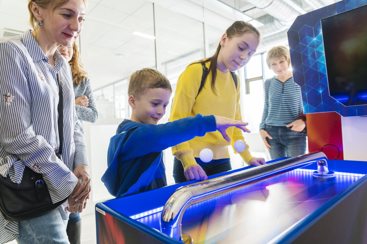 A boy is dropping a ball into an arcade machine while family members look on in a brightly lit indoor hall.