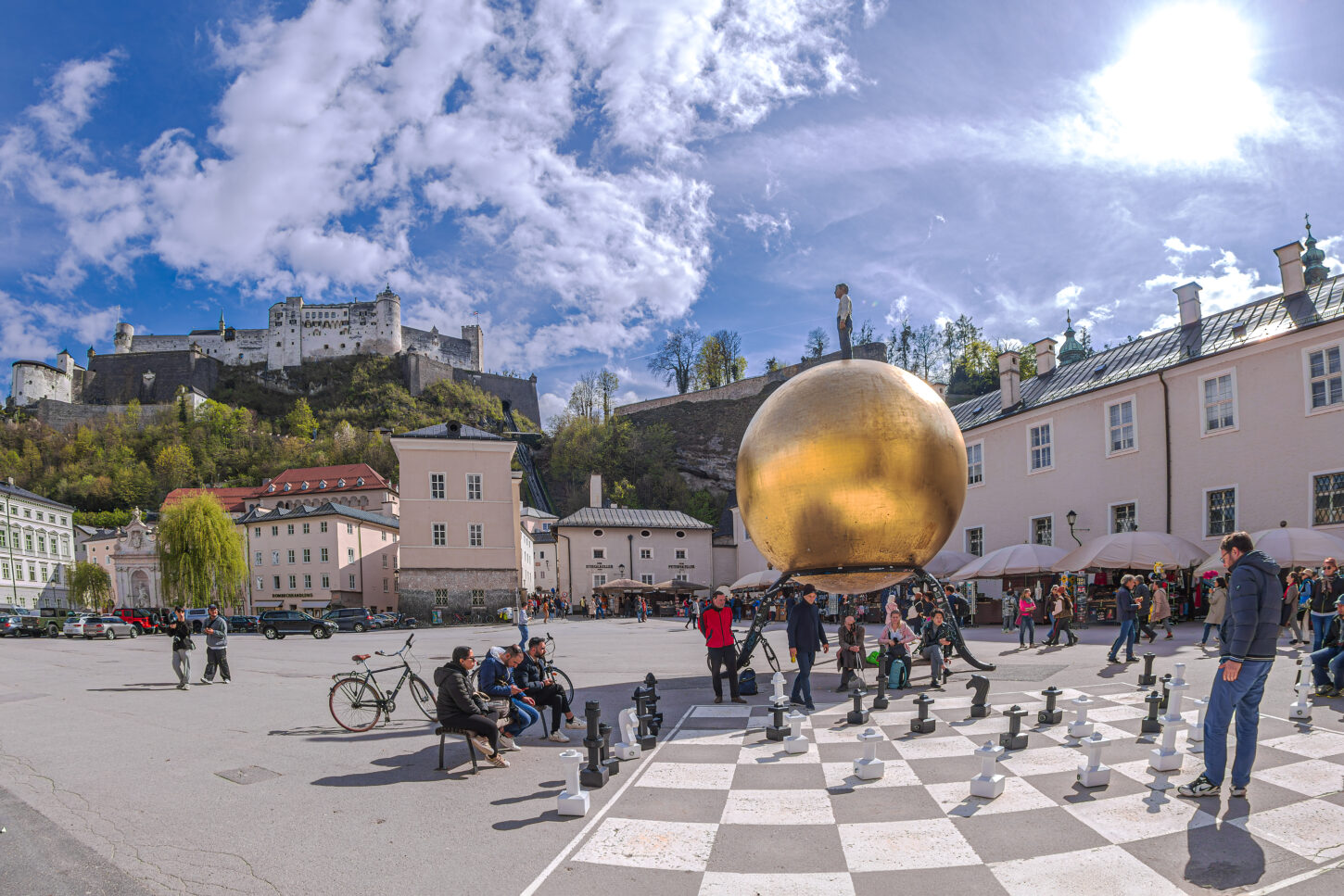 People playing a chess game drawn on asphalt in Kapitelplatz, near monument to Paul Fuerst "Sphere", a golden ball on which stands a small man. Castle Hohensalzburg in background.