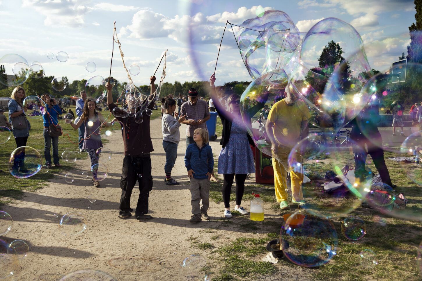 A group of people making giant soap bubbles on an early summer Sunday afternoon at Mauerpark, with the park's crowd scattered around them, some spectating.