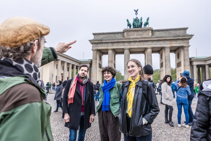 A group of people standing in front of the Brandenburg Gate in Berlin, with a tour guide in the foreground pointing at the monument.