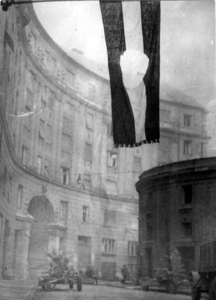 A black-and-white photograph from 1956 showing the Hungarian flag with a hole in it waving above some tanks in front of a large building in Budapest, Hungary.