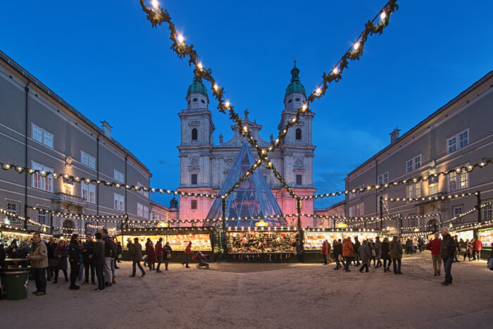 Christmas market at the Domplatz (Cathedral Square) in front of Salzburg Cathedral in twilight, with people walking around the square.