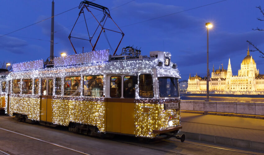 A brightly lit Christmas tram going on the street at night with the building of Hungarian Parliament alongside the Danube river in the background