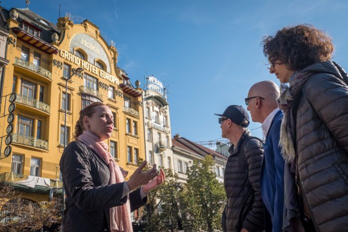 A tour guide talking to a small group of people in front of a yellow building on a bright sunny day