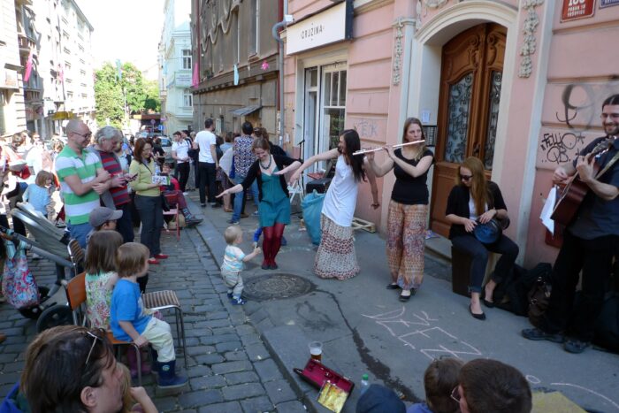 A group of people performing music on Krymska street in Prague on a sunny day, with a crowd of people watching.