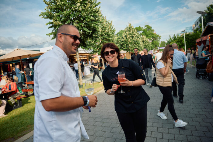 Two people with sunglasses holding wine glasses smiling at the camera, with people milling about looking at vendors, green leafy trees, and blue sky in he background