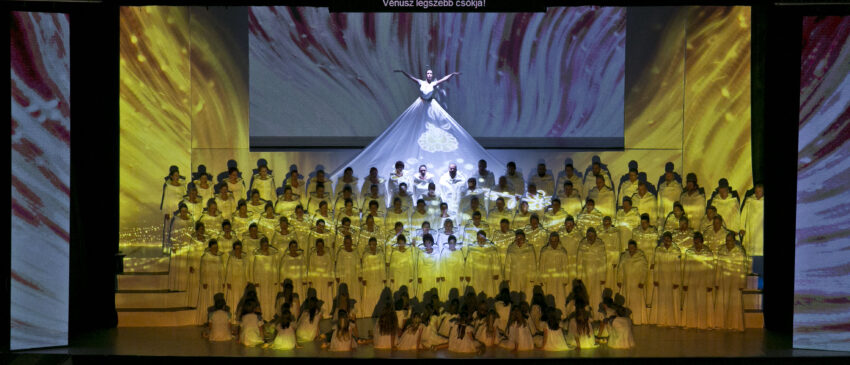 A choir singing on stage with a woman in a white dress in the top row, al bathed in yellow light