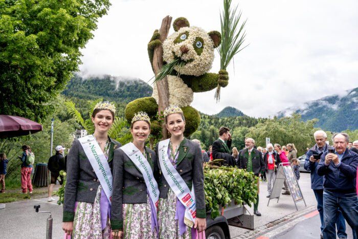 Three smiling women with narcissus crowns wearing sashes naming them Narcissus princess standing in front of a parade float with a panda made of plants