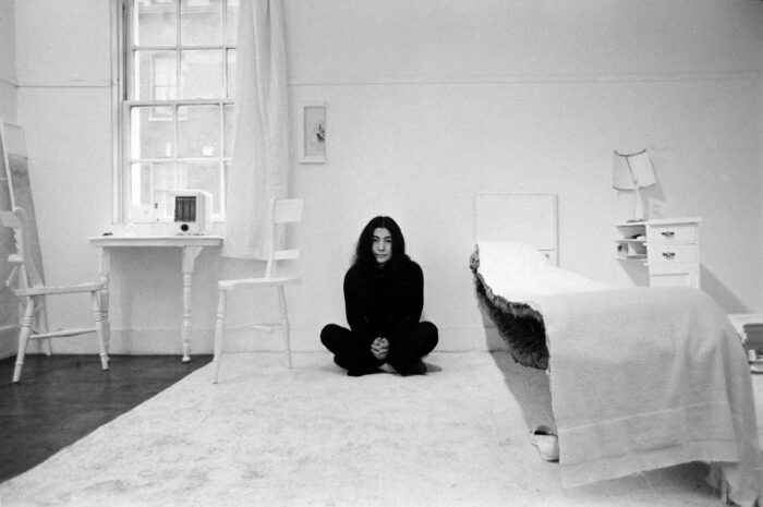 Black and white photo of Yoko Ono sitting in a room where all the furniture is cut in half, part of her piece Half a Room