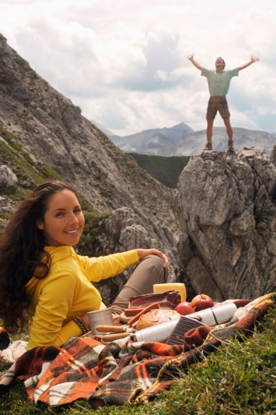 A young woman with dark hair lying on a picnic blanket and smiling at the camera in the foreground, while a man poses with triumph on a mountain peak in Salzkammergut, Austria, near Salzburg